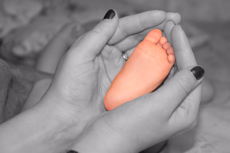 Zwei Hände halten einen winzigen Babyfuß, schwarzweiß, nur der Fuß ist farbig
