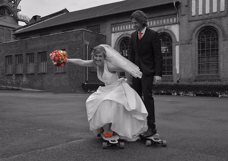 Braut auf einem Skateboard neben dem Bräutigam vor einem alten Backsteingebäude, schwarzweiß mit farbigem Brautstrauß