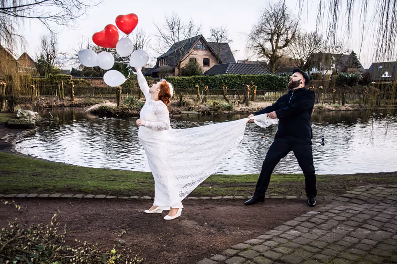 Braut mit roten Herzluftballons, der Bräutigam hält ihre Schleppe, dahinter ein Teich