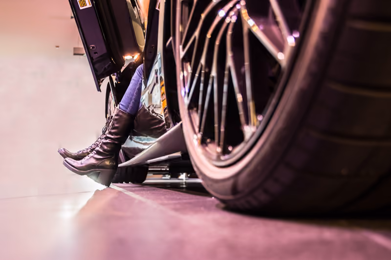 Frau in Stiefeln steigt aus einem Sportwagen, im Vordergrund die Felge