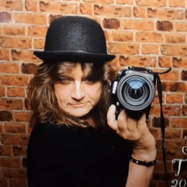 Susanne Bürger mit Hut und Kamera vor einer Backsteinwand